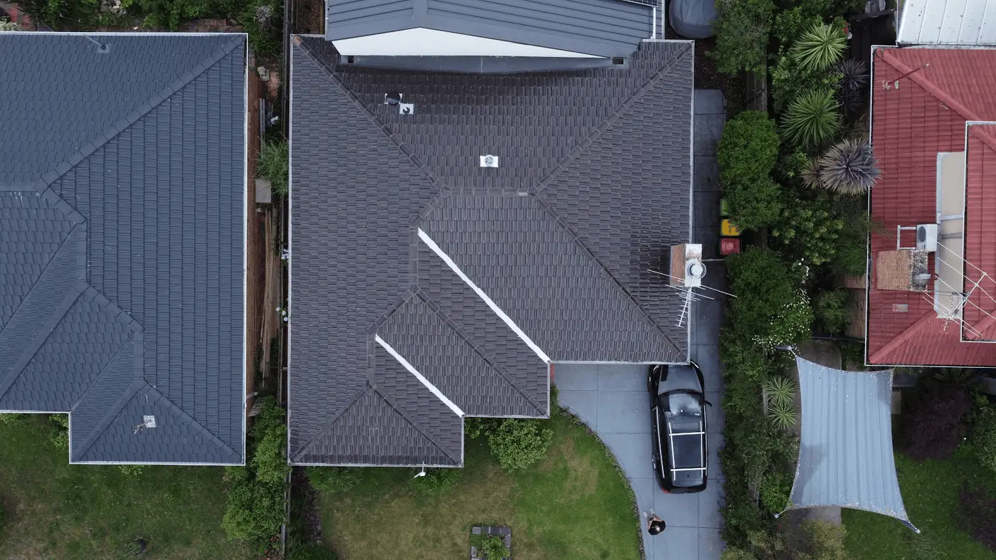 Aerial view of a house with a gray tiled roof, driveway, parked black car, green lawn, and surrounding vegetation. Neighboring houses are visible on either side.