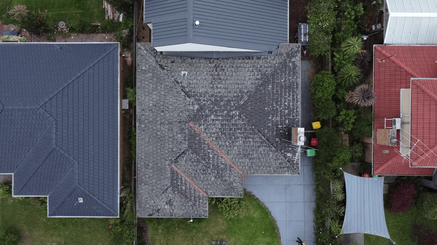 Aerial view of three neighboring houses with distinct roofs: grey shingles in the center, dark blue on the left, and red on the right. Driveway, green lawn, garbage bins, and trees are visible around the central house.