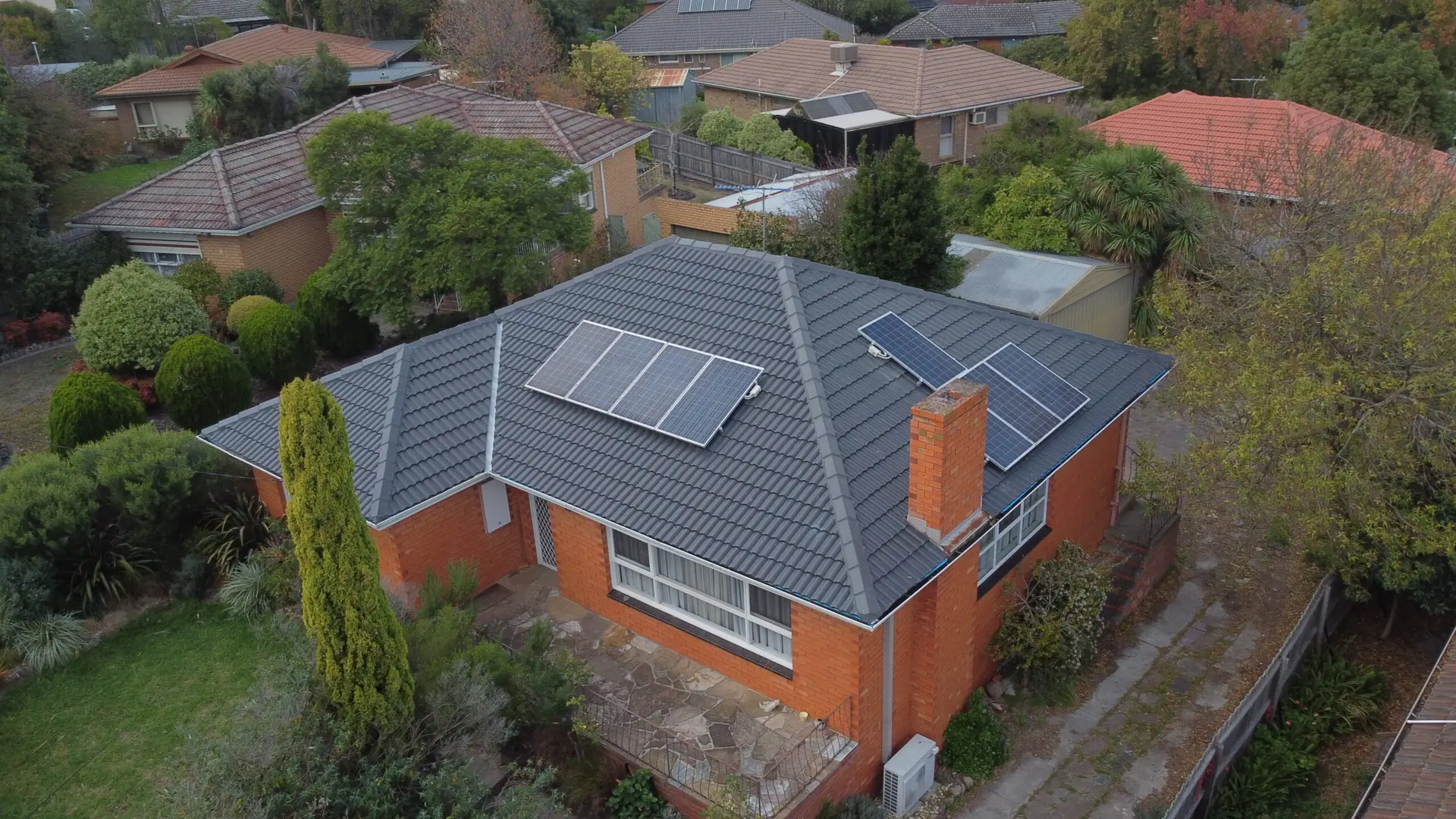 Aerial view of a red-brick house with a dark tiled roof equipped with solar panels, surrounded by greenery and neighboring houses in a suburban area.