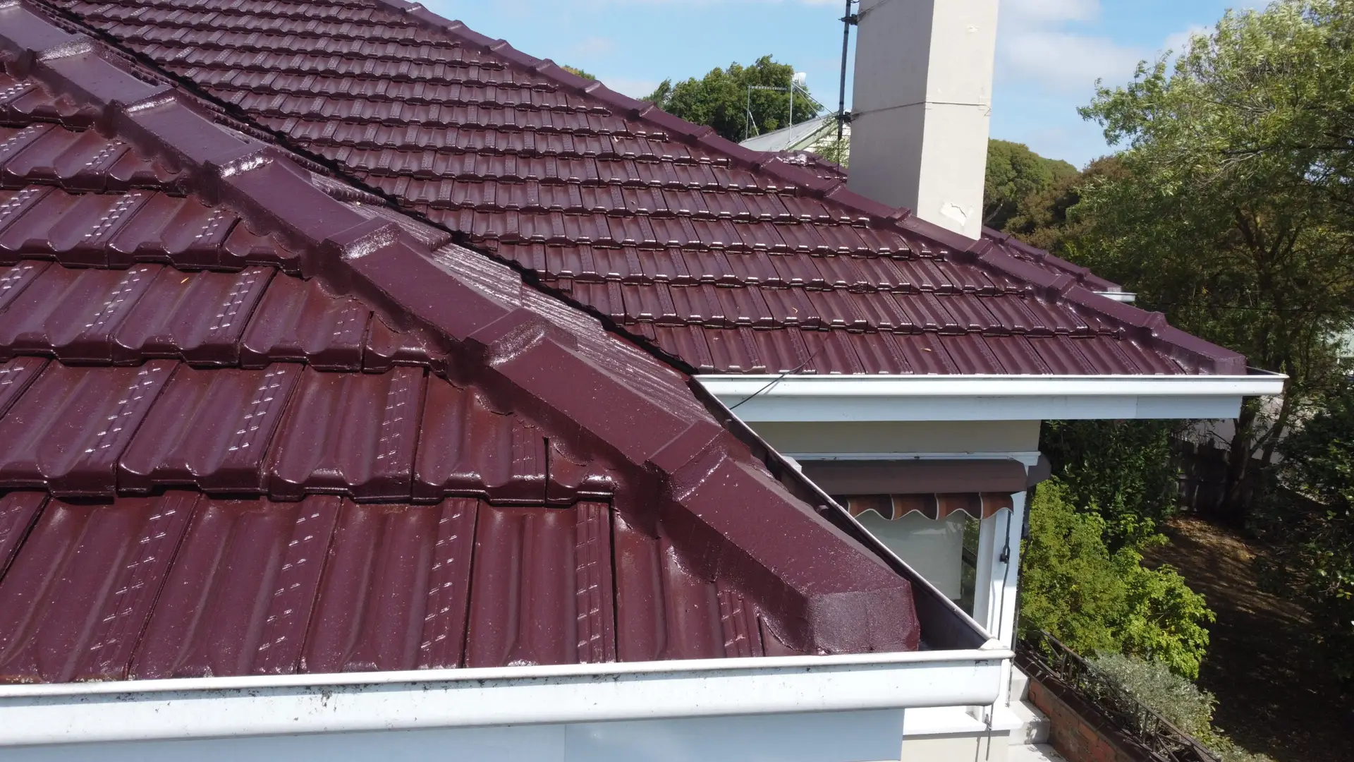 A close-up view of a house roof with shiny, maroon-colored metal tiles, white gutters, a chimney, and green trees in the background under a partly cloudy sky.
