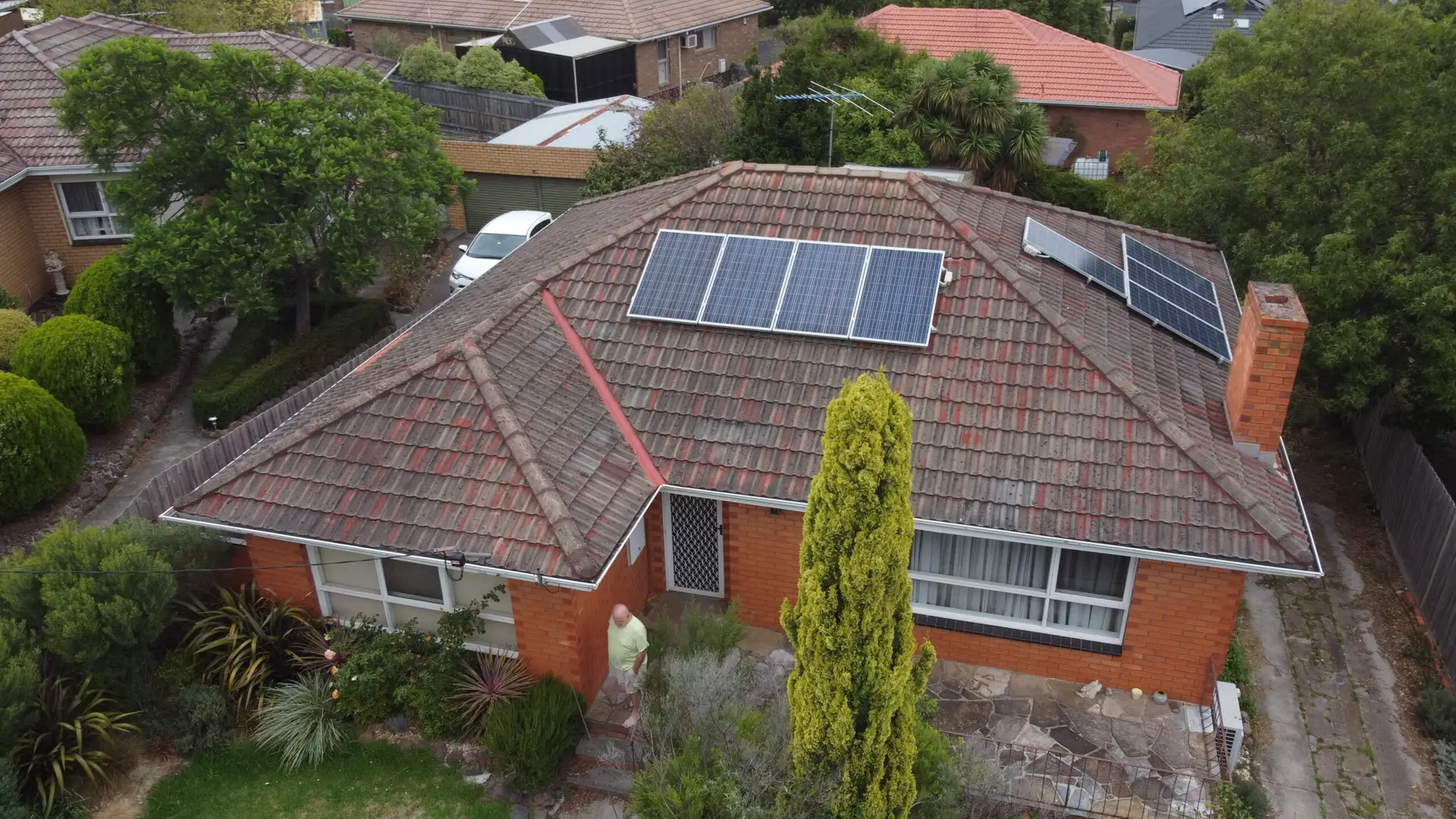 Aerial view of a single-story brick house with solar panels on the tiled roof, surrounded by trees, shrubs, and a garden path leading to the front door; neighboring houses are visible in the background.
