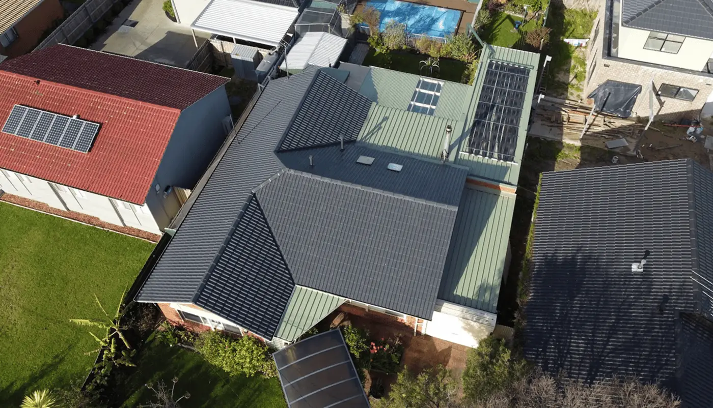 Aerial view of a house with multiple roof sections, including tiled and metal roofs. Solar panels are installed on one roof section, and the house is surrounded by gardens and neighboring homes.