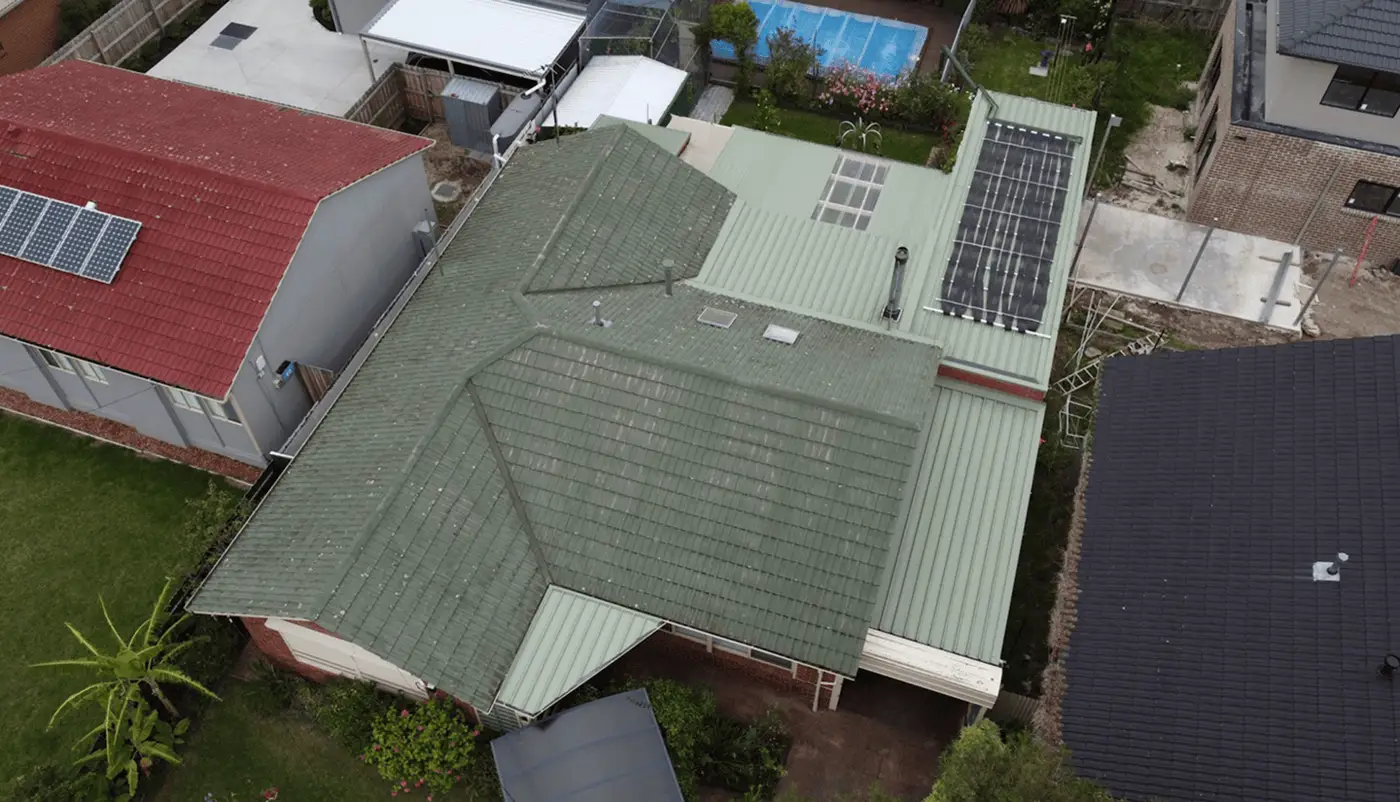 Aerial view of a house with a green tiled roof and solar panels, surrounded by neighboring homes, a backyard garden, and a blue swimming pool in the background.