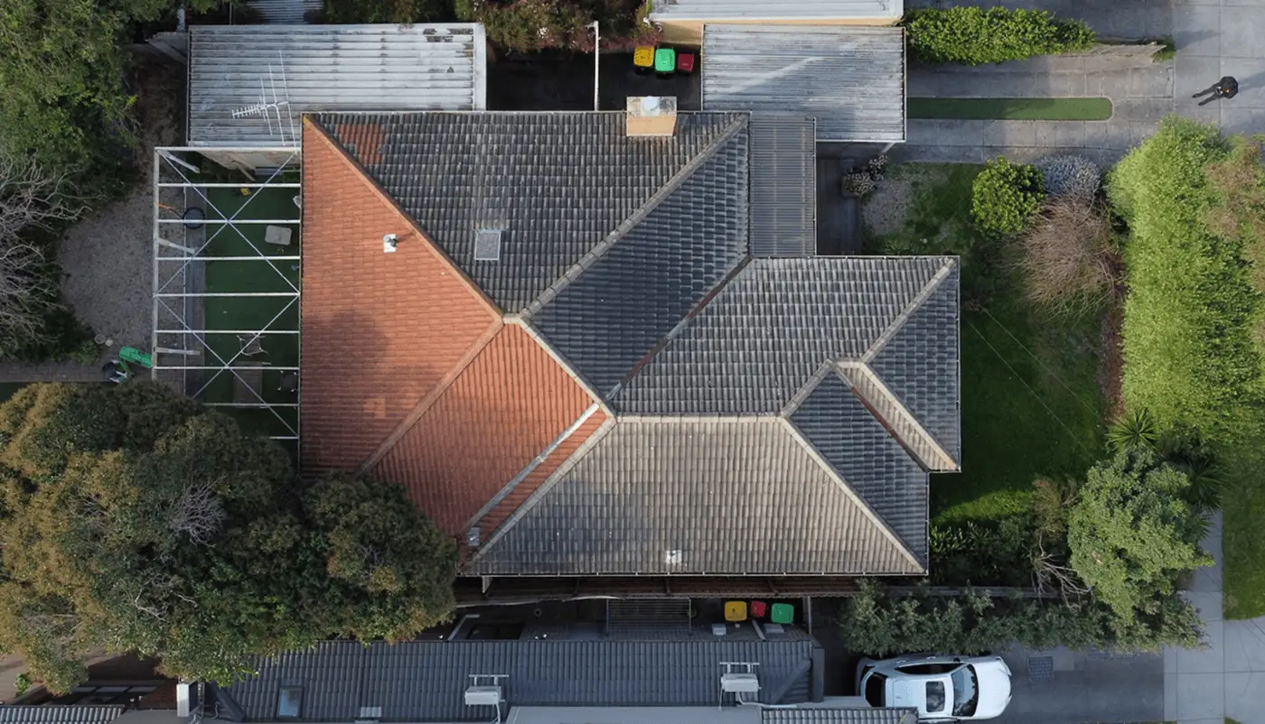 Aerial view of a house with a mix of dark and reddish roof tiles, surrounded by greenery, driveways, parked cars, and trash bins near the entrance.