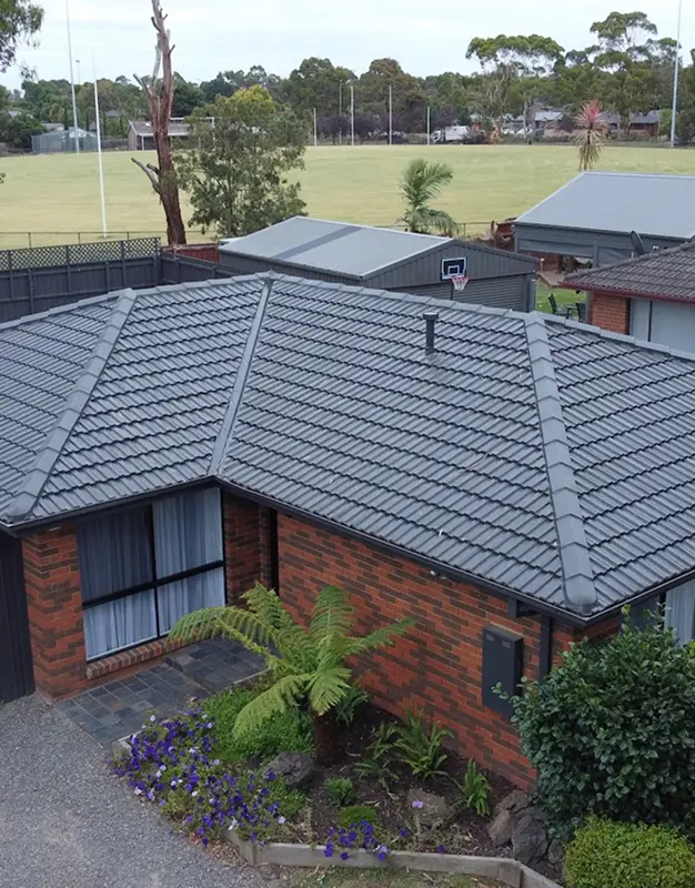 Single-story brick house with a grey tiled roof installed by a trusted roofer, surrounded by greenery and a small garden. A fenced sports field and trees are visible in the background under an overcast sky.