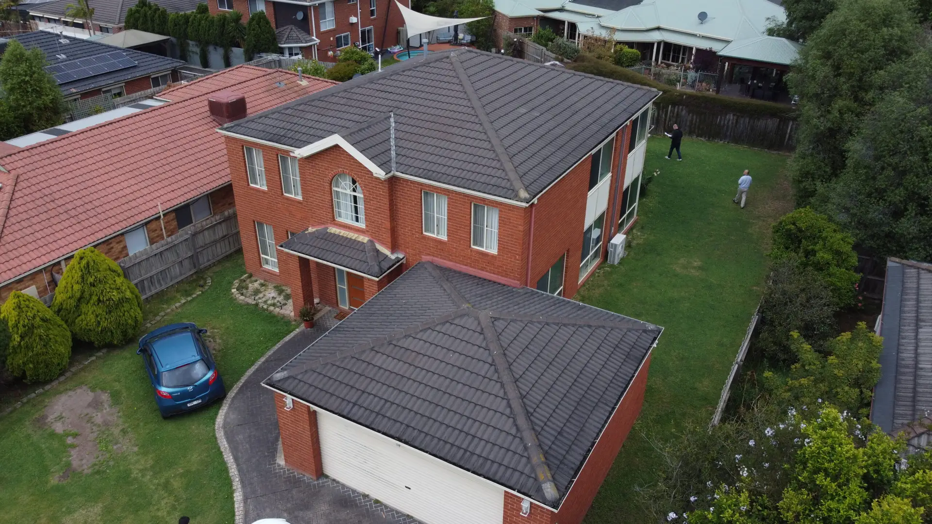 Aerial view of a large two-story brick house with a gray tiled roof, a driveway with a blue car, a front yard with bushes, and three people standing in the backyard.