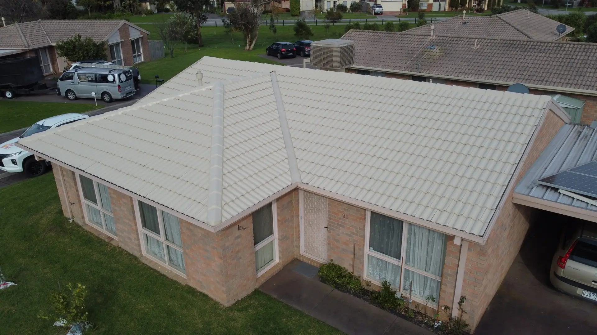 Aerial view of a single-story brick house with a light-colored tiled roof, large windows, and a small green lawn, situated in a suburban neighborhood with nearby houses and parked cars.