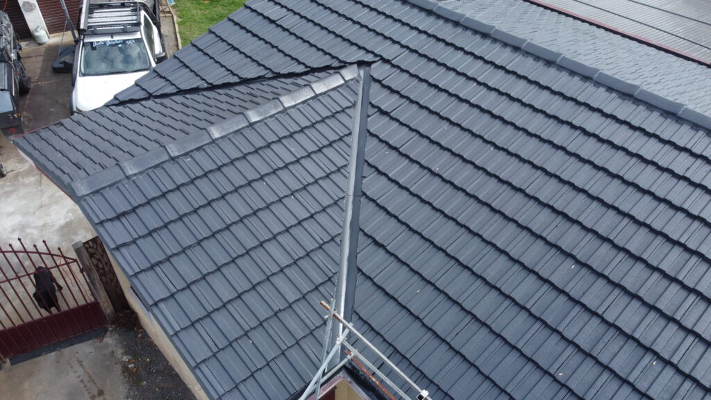 Close-up view of a freshly repaired dark grey tile roof in Croydon, showing clean ridges, restored valley flashing, and a well-maintained finish beside a driveway and a red metal gate.